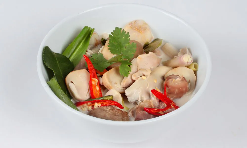 Thai coconut chicken soup in white bowl, filled with chicken pieces, mushrooms, red chili slices, cilantro, lemongrass and kaffir lime leaves.