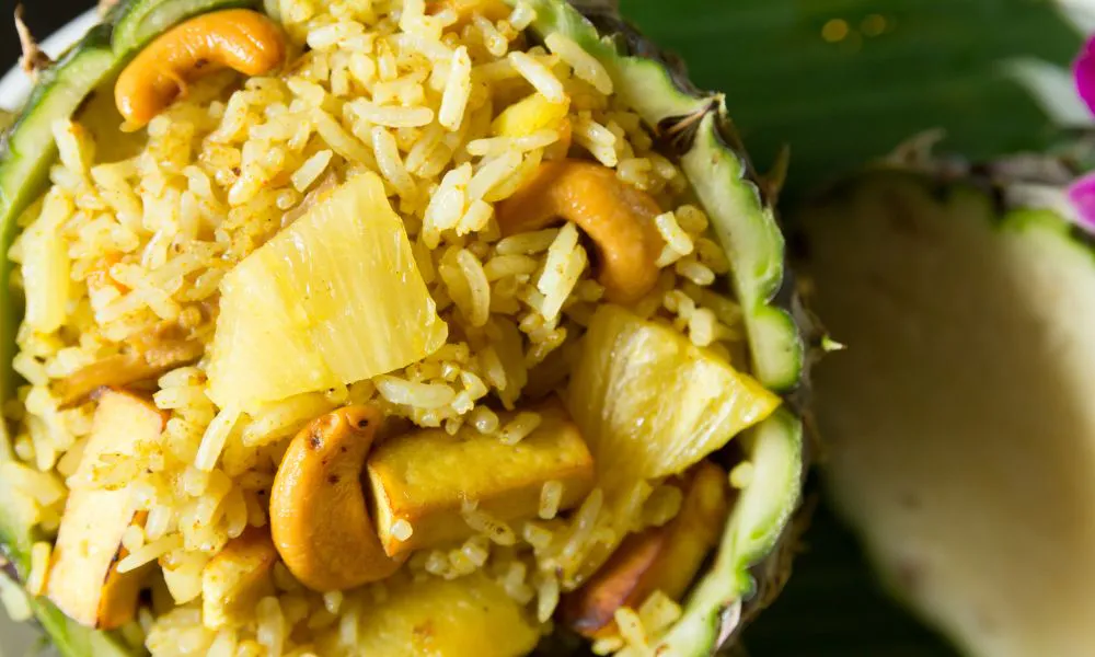 Top‑down close‑up of pineapple fried rice served inside hollowed pineapple shell, mixed with cashew nuts, pineapple chunks and tofu pieces.